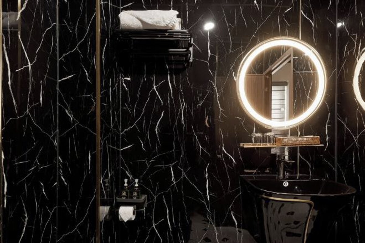 Un baño con pared de mármol negro, un espejo redondo con halo de luz y un lavamanos negro.