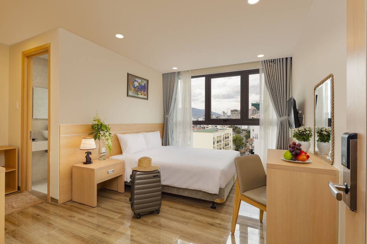 A hotel room with a bed, bedside table, lamp, suitcase, desk, chair, and a view of the city through the window.
