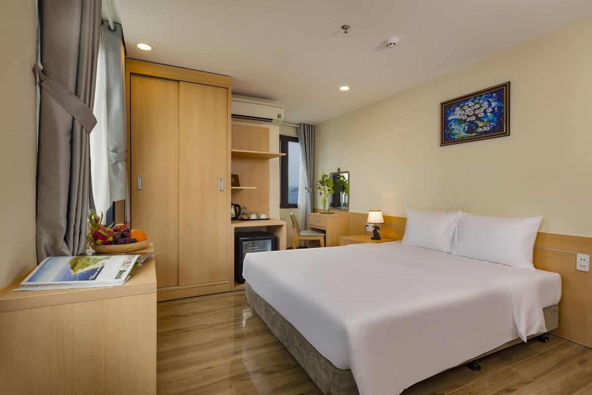 A hotel room with a bed, a desk with a fruit bowl, a small fridge, a mirror, and a painting on the wall.