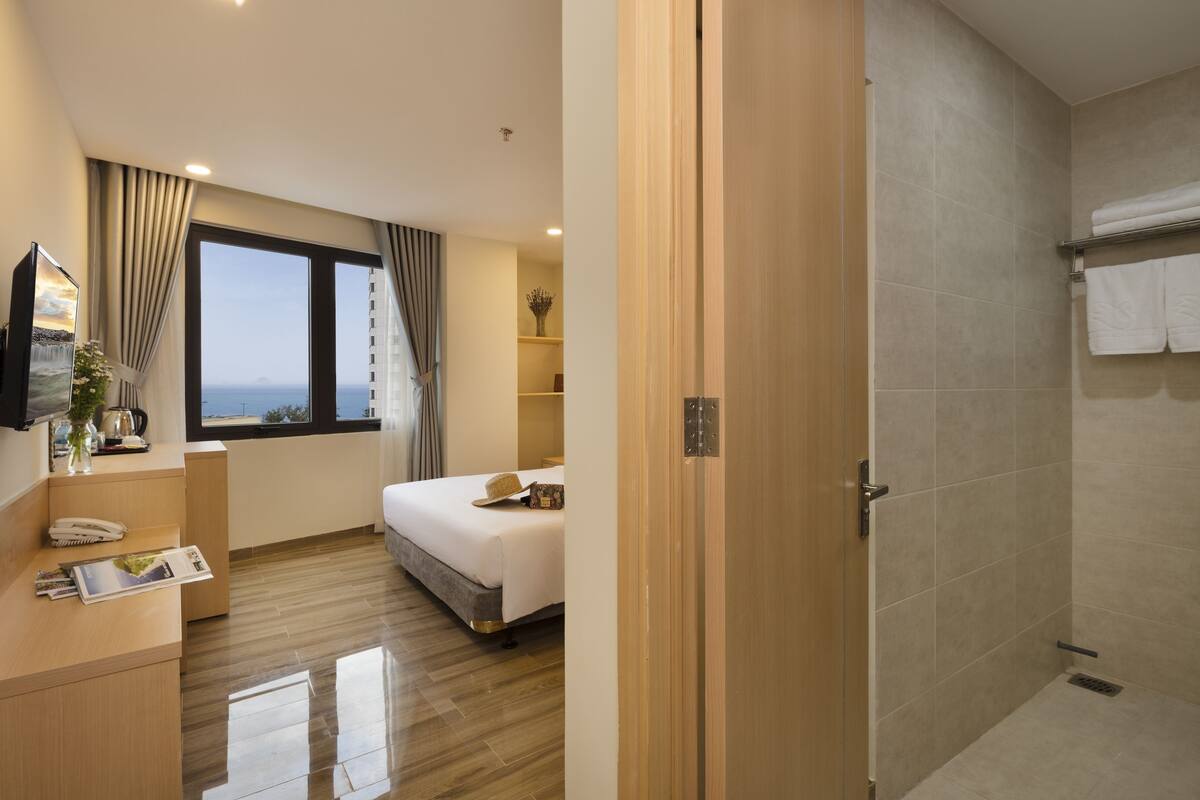 A hotel room with a bed, a desk with a telephone and a magazine, a window with curtains, and a shower area with a towel rack.