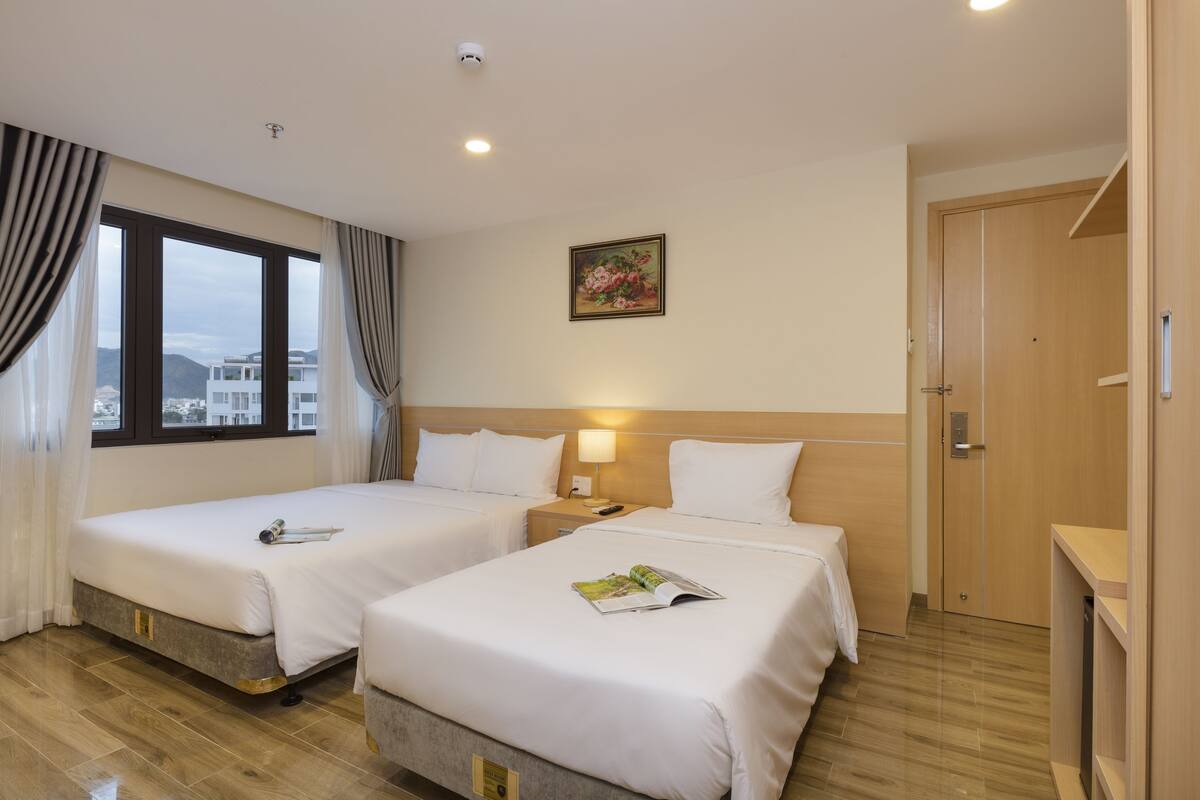 A hotel room with two single beds, a wooden wardrobe, a bedside table with a lamp, and a window with curtains.