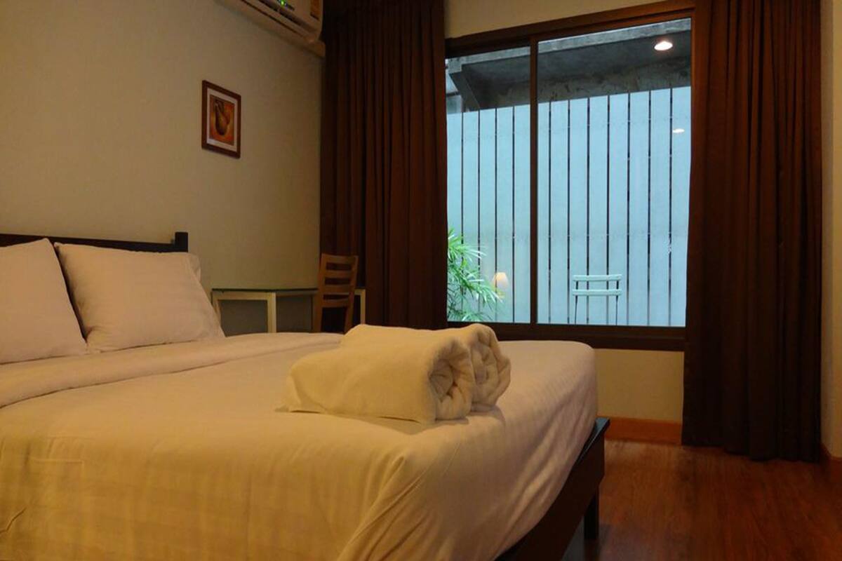 Una cama bien hecha con una toalla doblada, un cabecero de madera, una mesita y una ventana con cortinas.