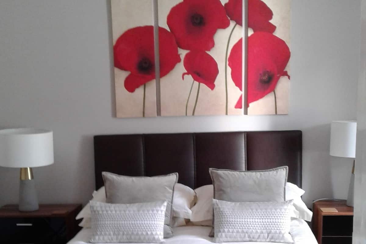 Ein ordentlich bezogenes Bett mit einer roten Decke, ein großes Gemälde von roten Blumen an der Wand und zwei Nachttischlampen.