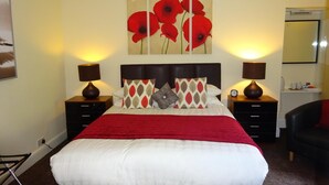 A bedroom with a large bed, two bedside tables with lamps, a wall-mounted artwork of red flowers, and a small bedside table with a clock. - Adelphi Villa (Oban)