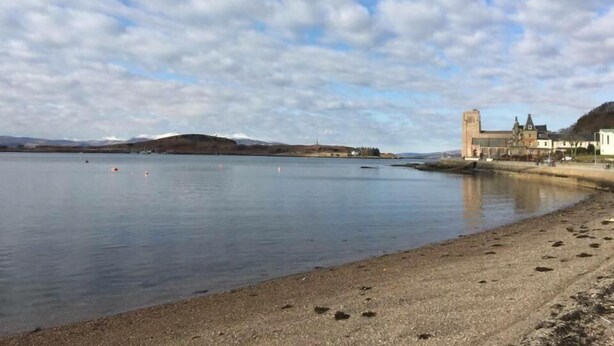 A coastal scene with a calm sea, a sandy beach, and a historic building with a tower. - Adelphi Villa (Oban)