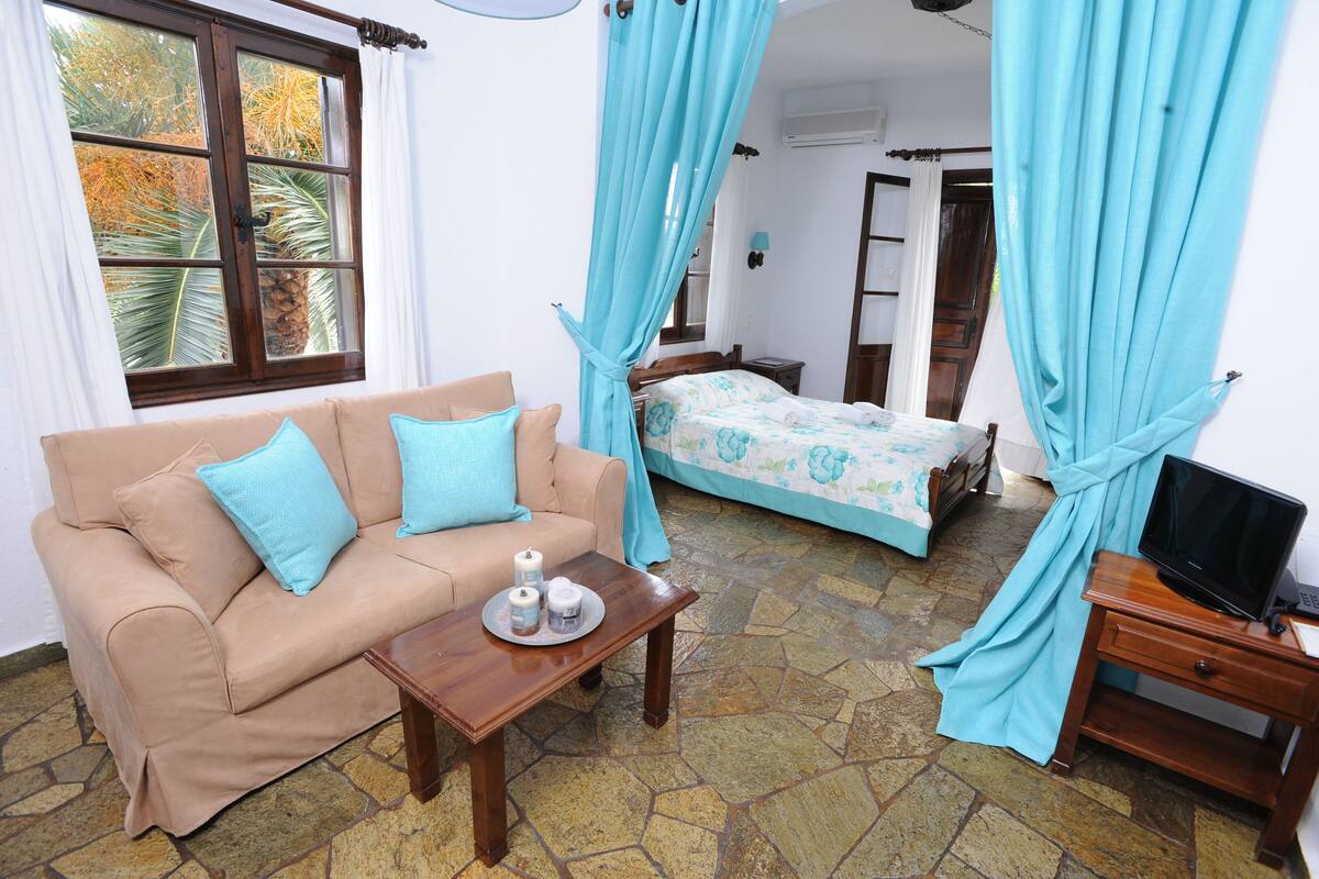 Una habitación con sofá color beige, cama con ropa de cama estampada, suelo de baldosas de piedra y televisor sobre un soporte de madera.