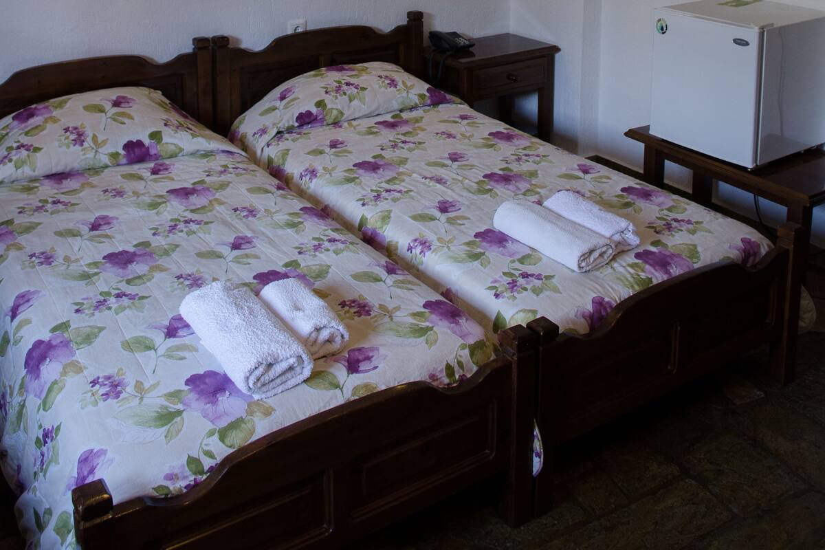 Habitación de hotel con dos camas, cada una con ropa de cama y toallas con motivos florales, un escritorio pequeño, un refrigerador y lámparas de pared.