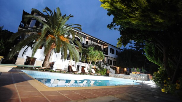 A poolside area with lounge chairs, palm trees, and a building with balconies. - Elli Hotel (Skopelos)