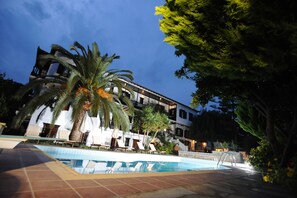 A poolside area with lounge chairs, palm trees, and a building with balconies. - Elli Hotel (Skopelos)