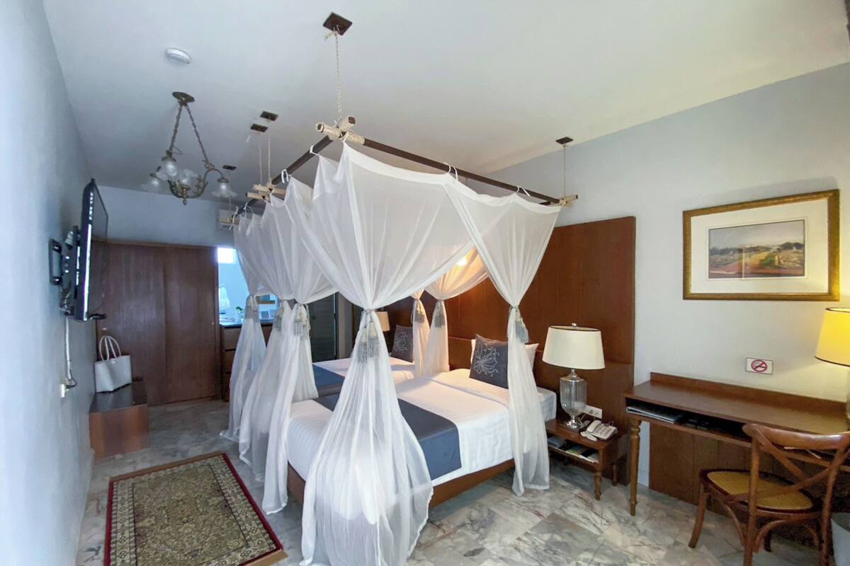 A hotel room with a four-poster bed, a desk, a chair, a nightstand, a lamp, and a small rug.