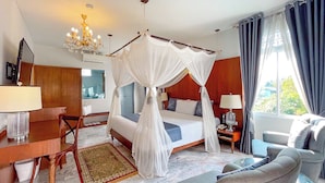 Habitación de hotel con cama con dosel, un escritorio con lámpara, una silla, un sofá y una mesita de noche con lámpara.
