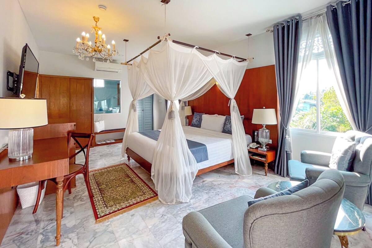 A hotel room with a four-poster bed, a desk with a lamp, a chair, a sofa, and a nightstand with a lamp.