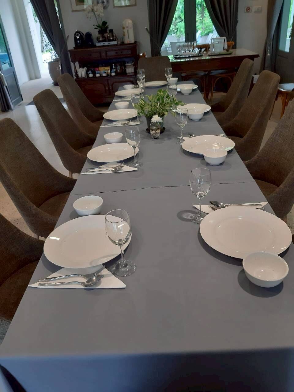 A dining table set for six with white plates, glasses, and cutlery, surrounded by brown chairs.