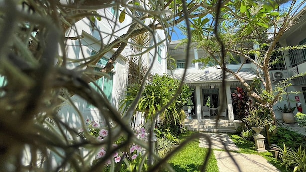 Un patio con un edificio blanco, una zona de estar cubierta y diversas plantas.