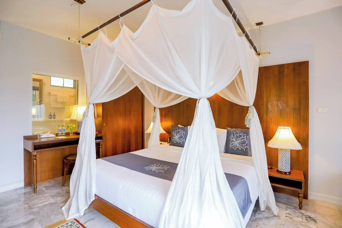 A four-poster canopy bed with a wooden headboard, a bedside table with a lamp, and a view of a bathroom with a mirror and towel rack.