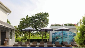 Zona de piscina con sillas de descanso y sombrillas, un mural y un árbol.