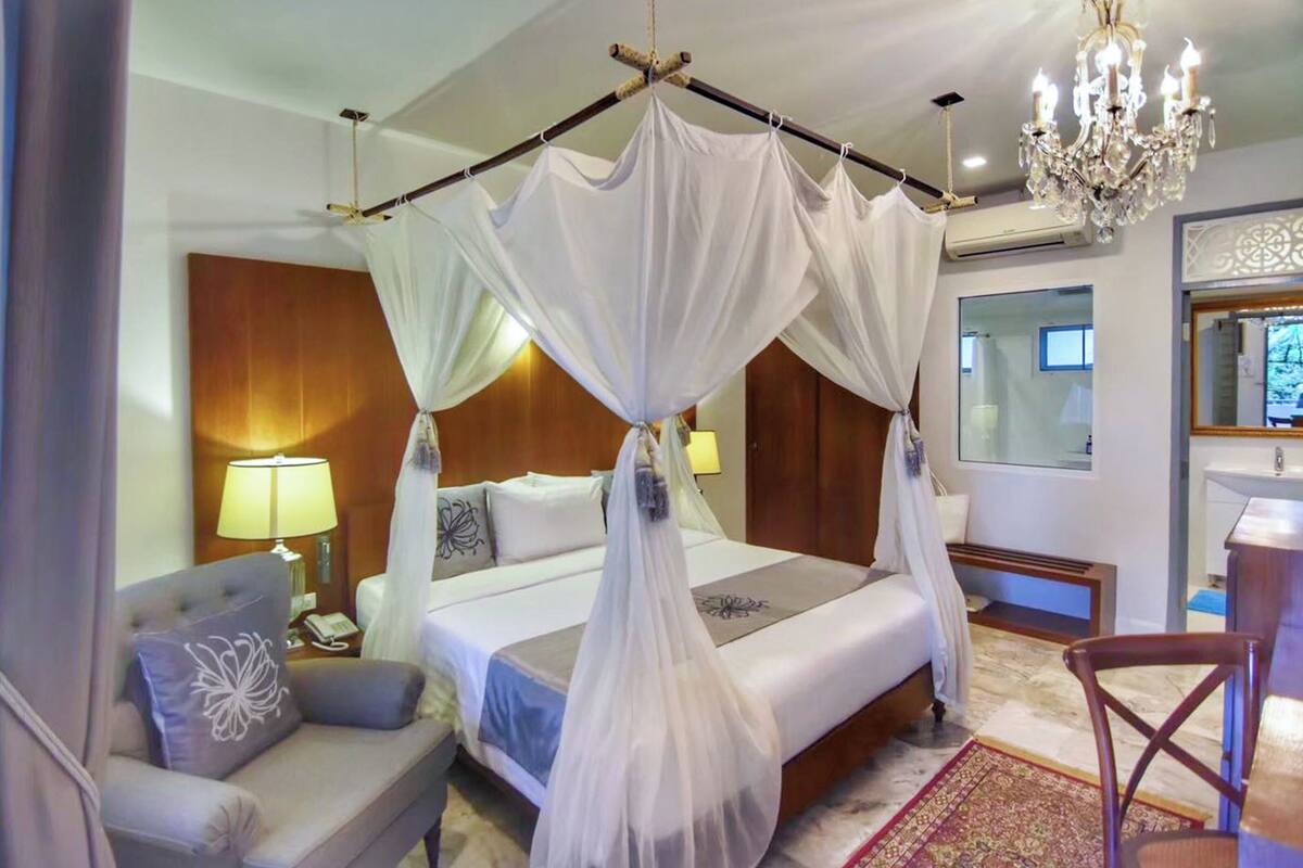 A hotel room with a four-poster bed, a chandelier, a bedside table with a lamp, a sofa, and a wooden chair.