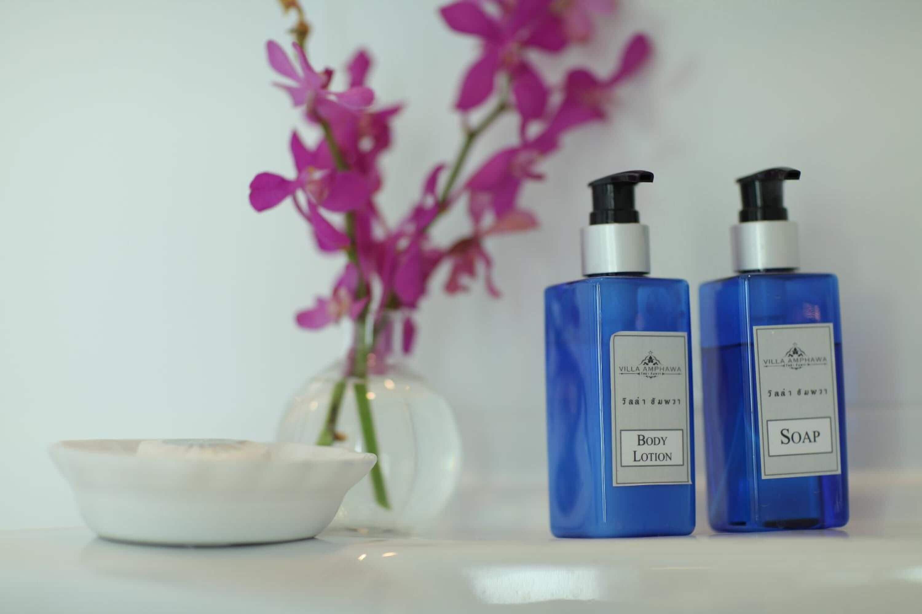 Two blue bottles labeled 'Body Lotion' and 'Soap' next to a white bowl and a vase with pink flowers.
