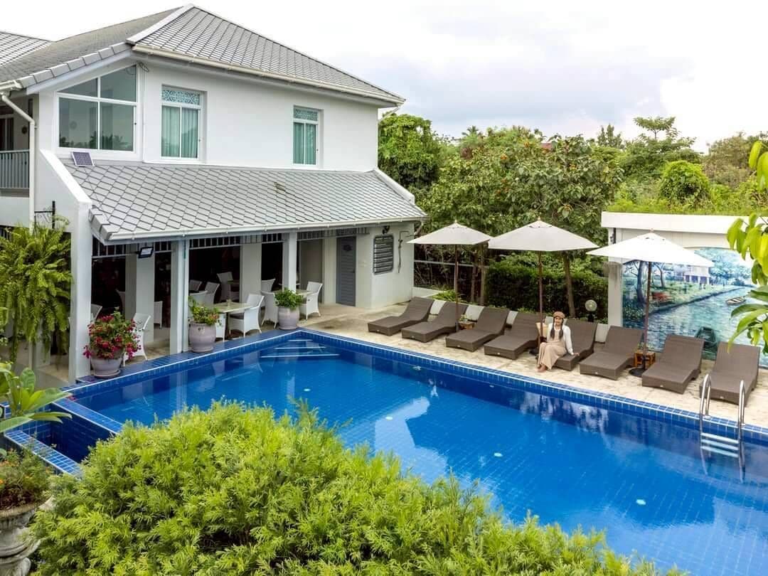 Una piscina con sillas de descanso y sombrillas, rodeada de vegetación y un edificio blanco.