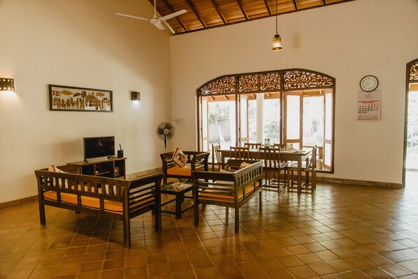 A spacious room with a high ceiling, a ceiling fan, a large window with decorative woodwork, and a wall-mounted clock.