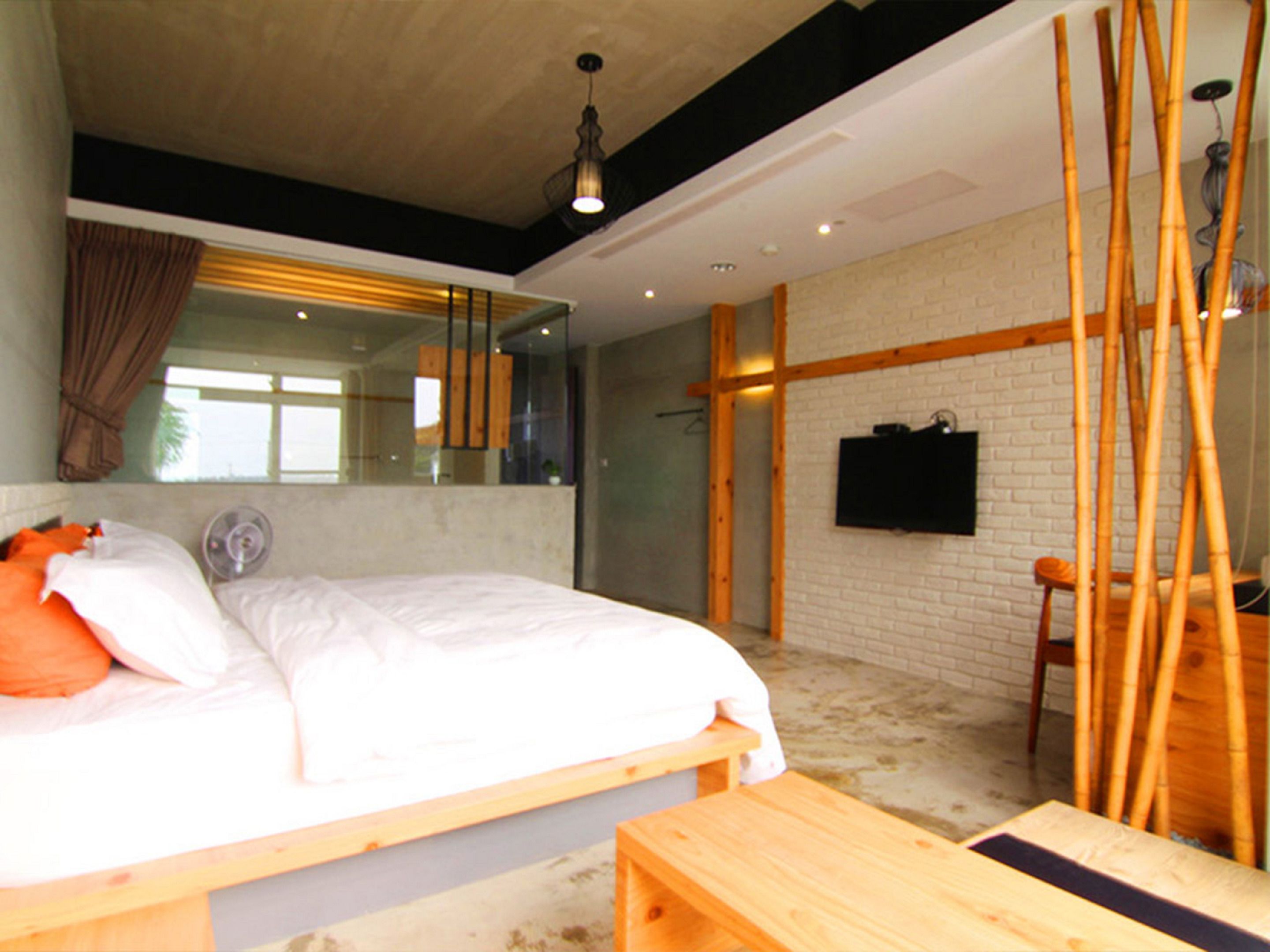 A modern bedroom with a large bed, a wooden bench, a TV mounted on a brick wall, and a large window with curtains.