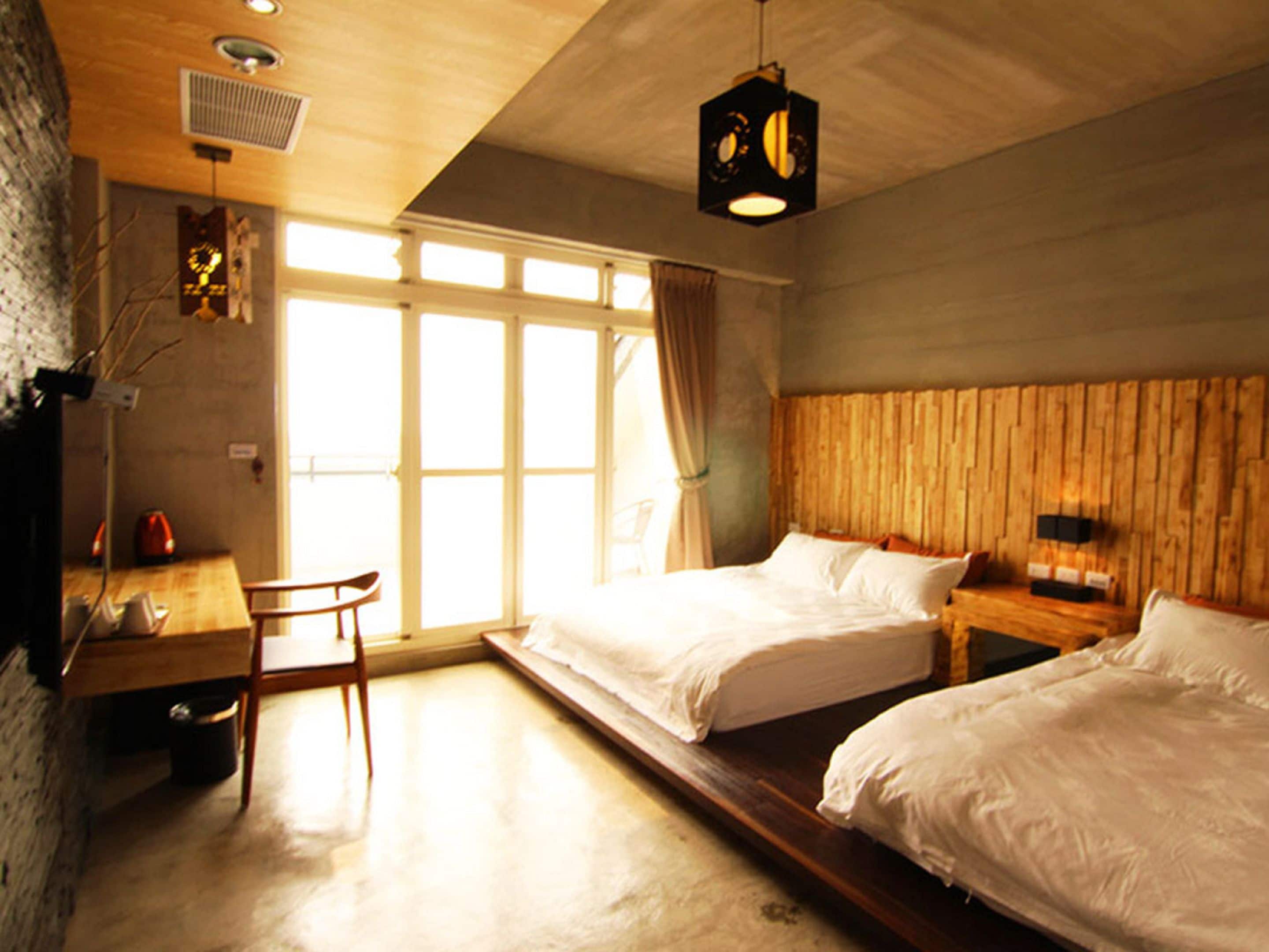 A room with two beds, a desk, and a chair. There is a large window with curtains, a wooden headboard, and a hanging light fixture.