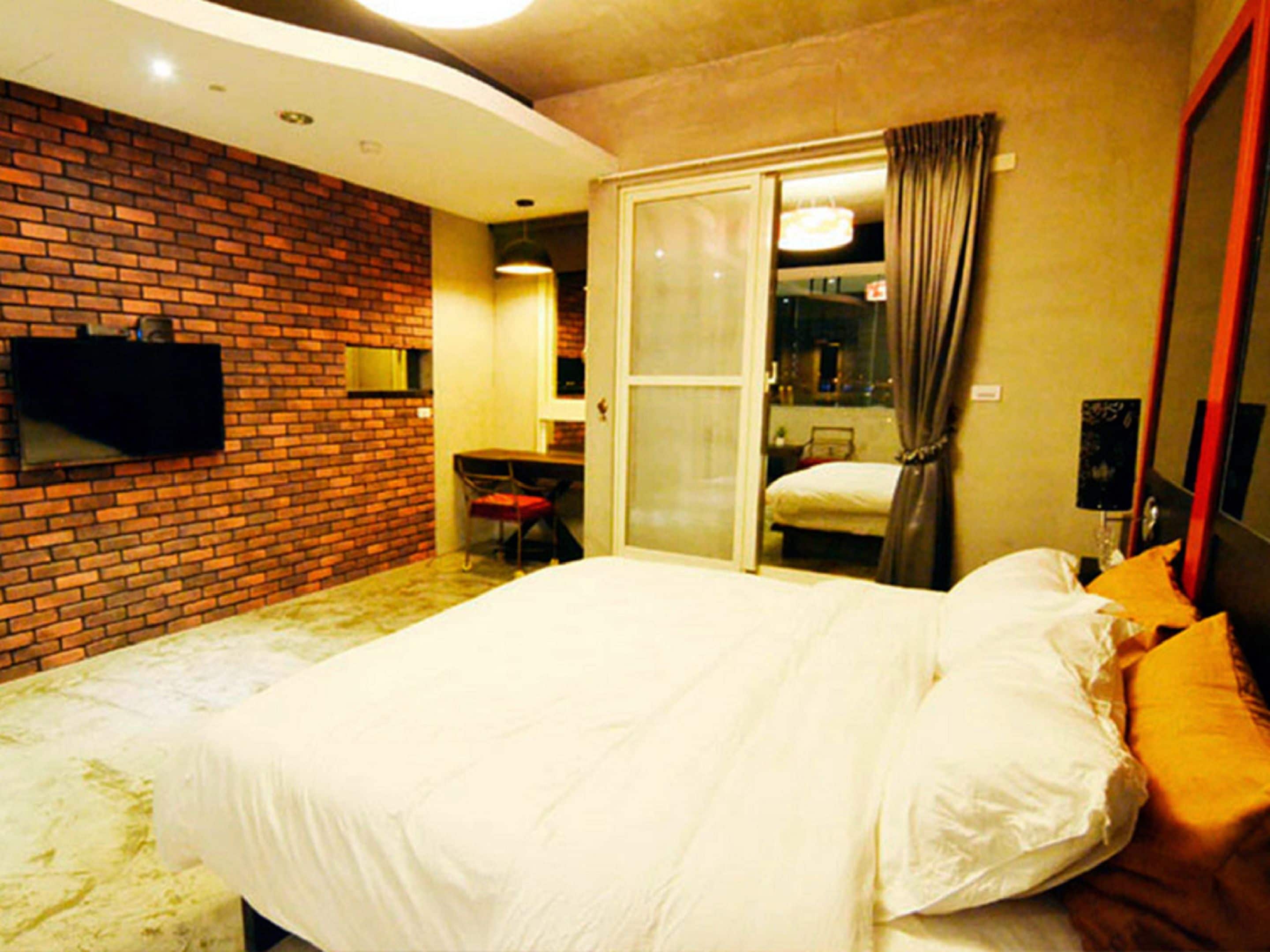 A hotel room with a brick wall, a flat-screen TV mounted above a desk, a bed with white linens, and a sliding glass door leading to a bathroom.