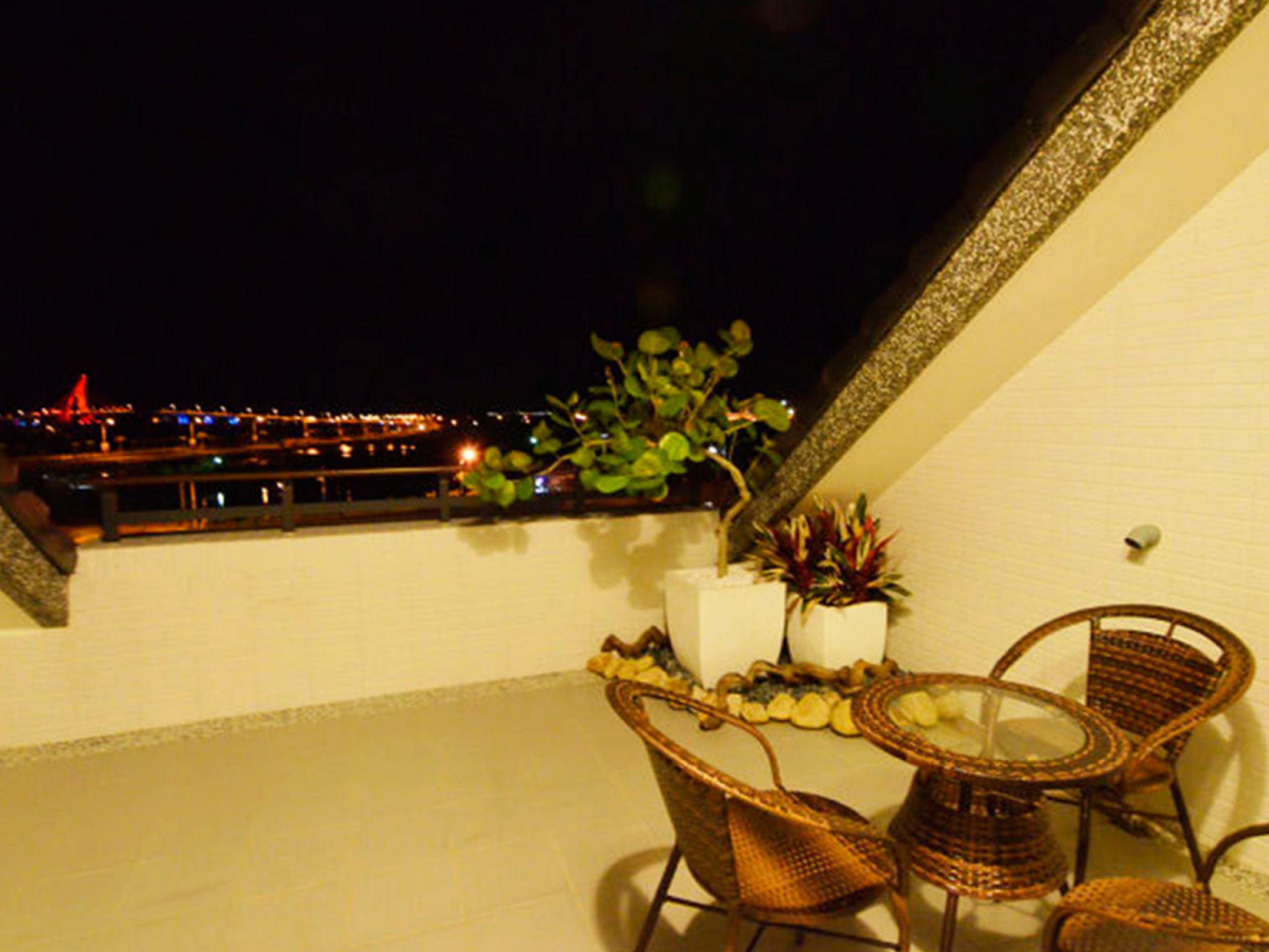 Ein Balkon mit Korbstühlen, einem kleinen Tisch, Blumentöpfen und Blick auf eine beleuchtete Stadtlandschaft bei Nacht.