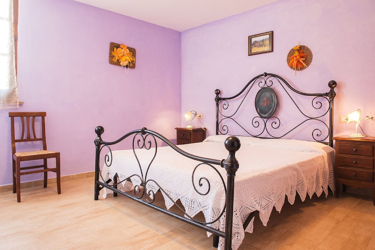 A bedroom with a bed, a chair, a nightstand, and a wall with two decorative items.