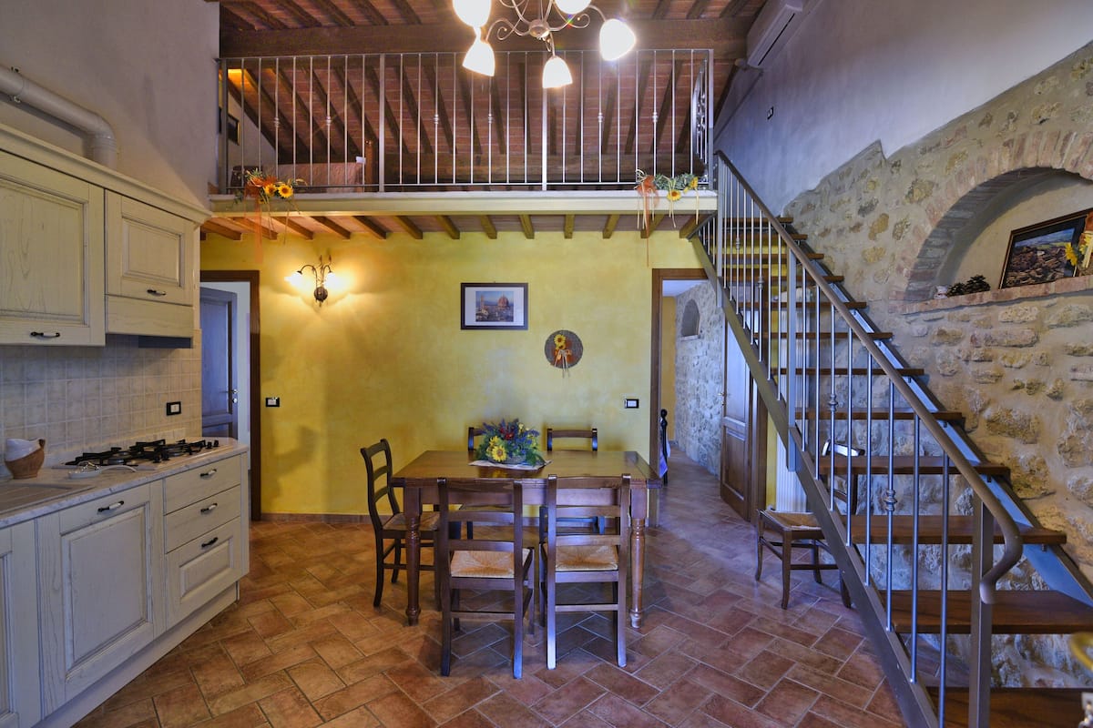 A dining area with a wooden table and chairs, a kitchen with cabinets, and a stone staircase.