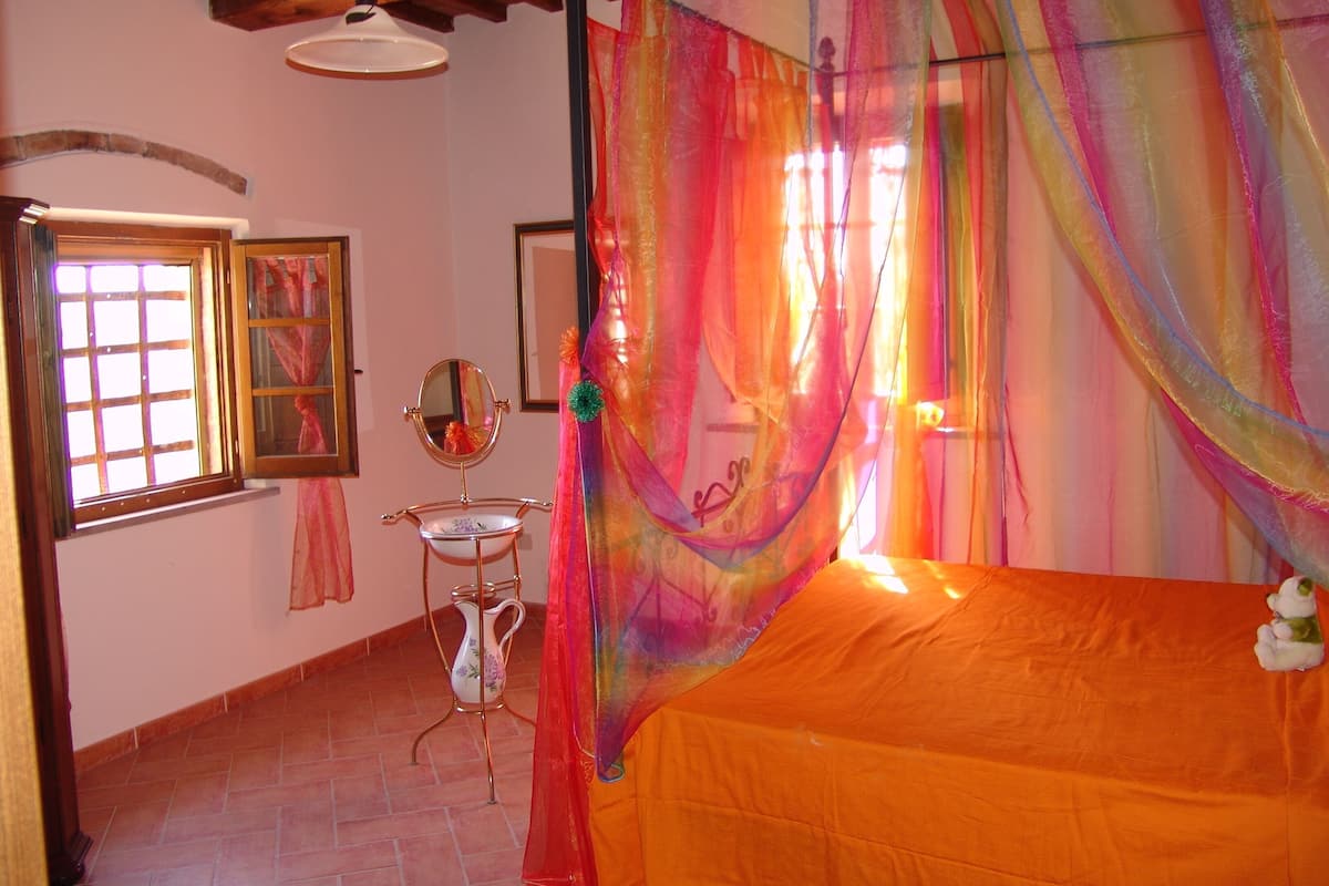A bedroom with a canopy bed, a window with curtains, a mirror, and a pitcher on a stand.