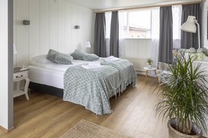 A bedroom with two beds, a bedside table, a lamp, a small table with a plant, and a large window with curtains.