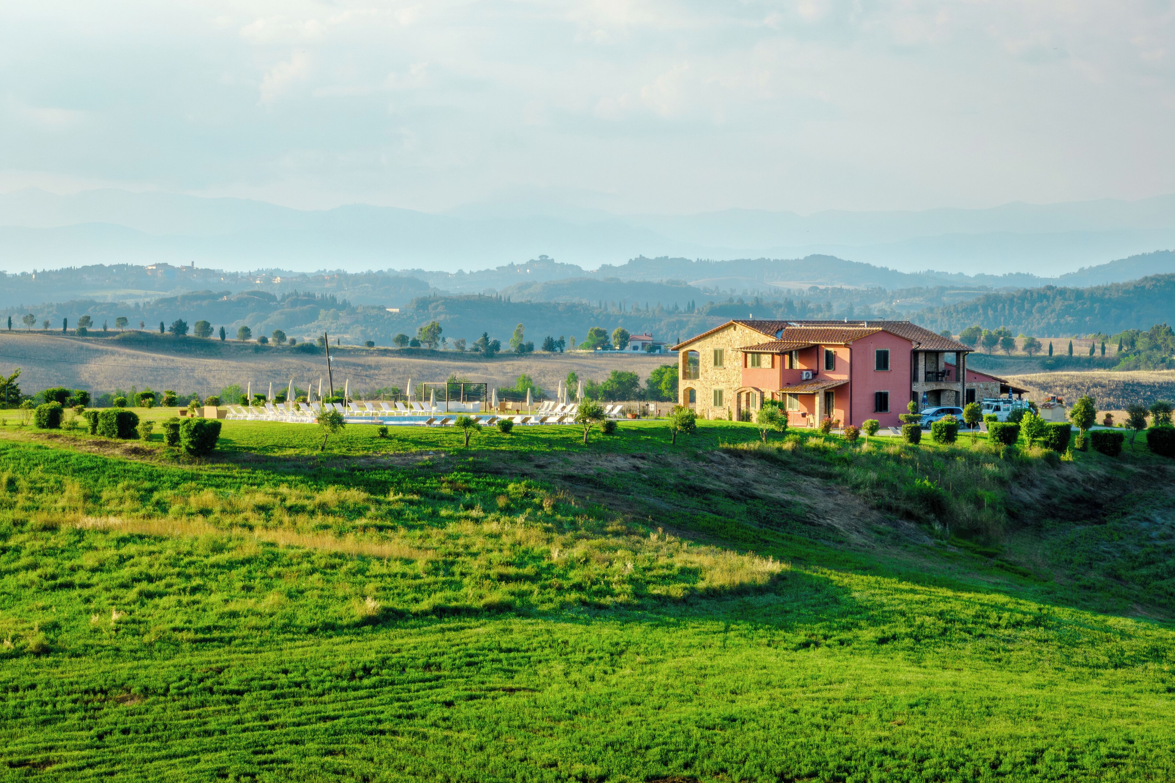 A house with a red roof and a balcony, surrounded by green fields and hills in the background.
