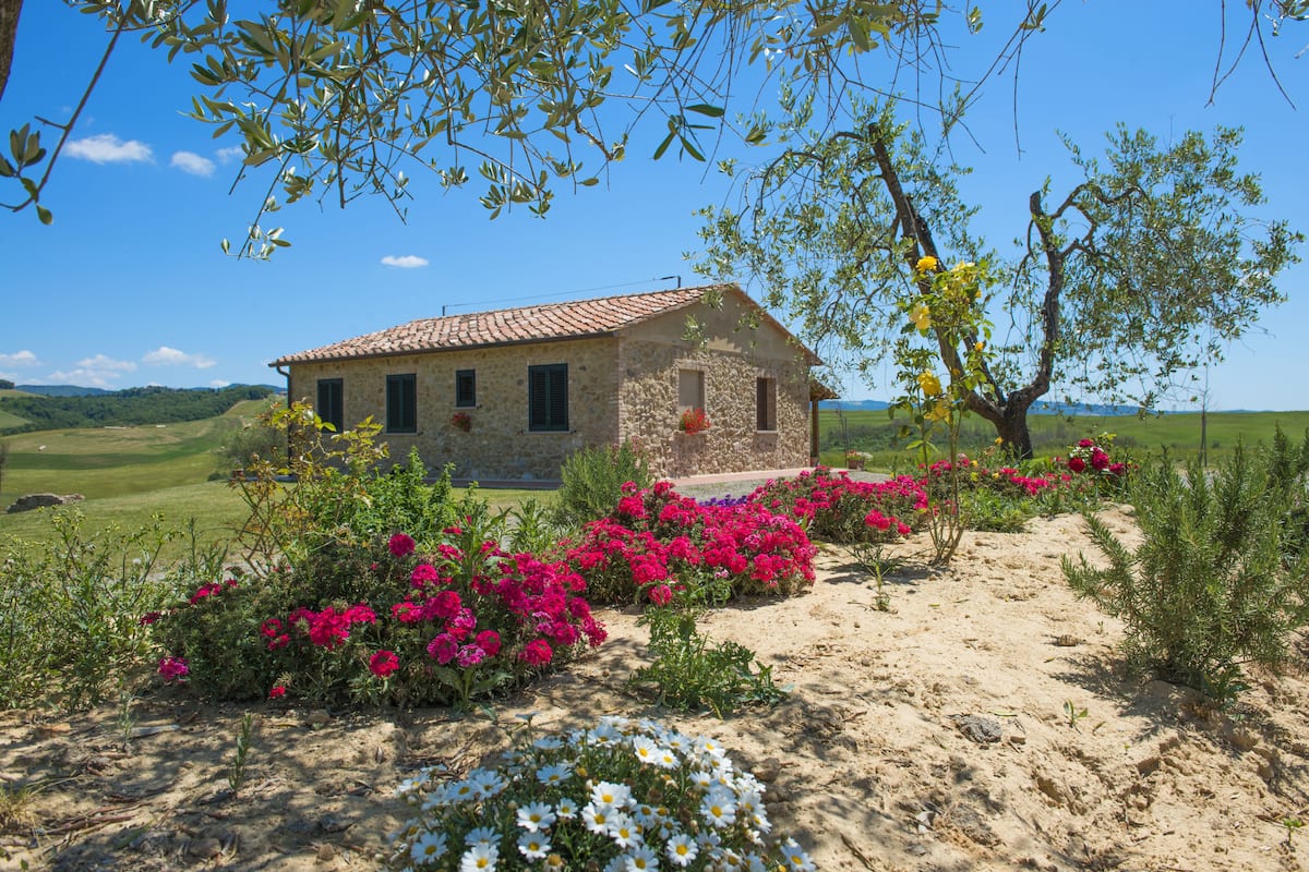 Uma casa de pedra com telhado de cerâmica, rodeada por um jardim com flores rosa e brancas, e um céu azul límpido.