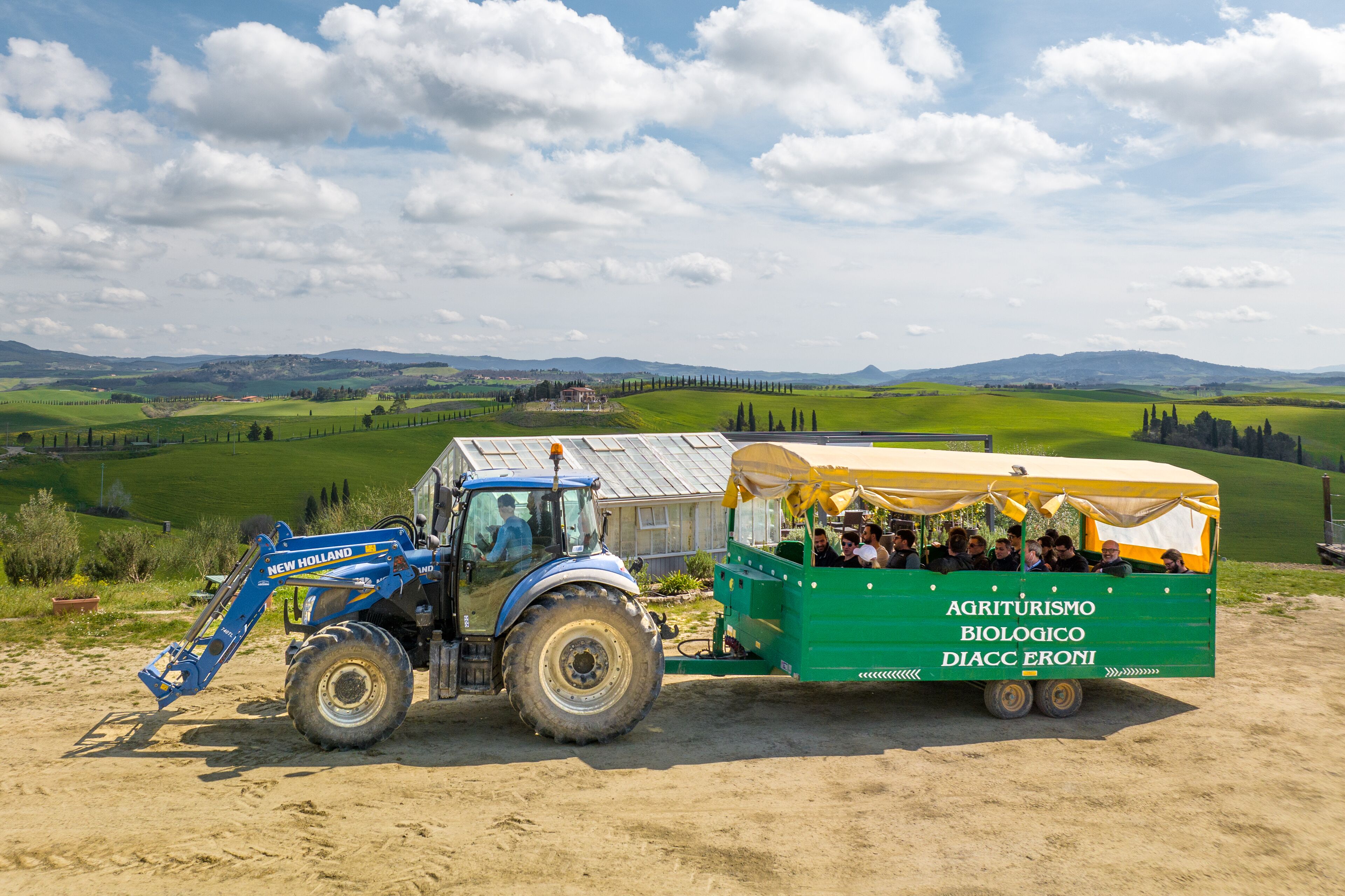 En blå traktor drar ett grönt släp med ett gult tak där det står ”AGRITURISMO BIOLOGICO DIACC ERONI”.