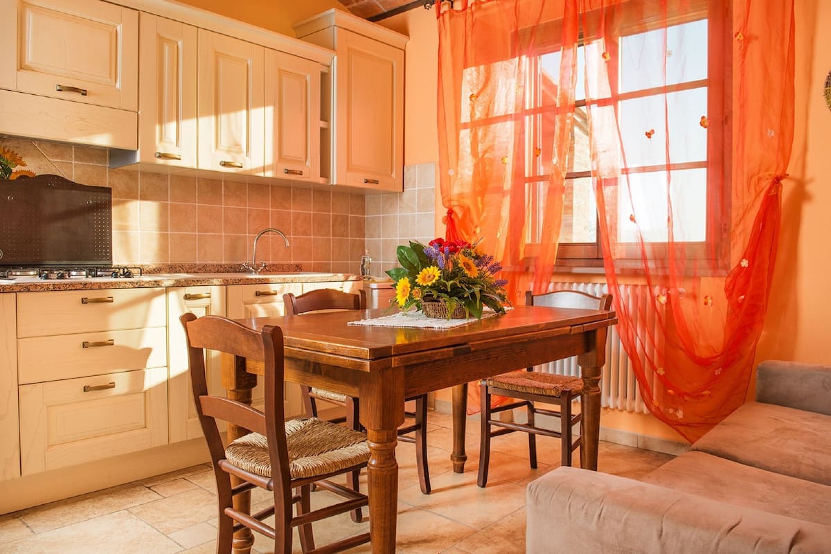 Uma cozinha com armários de madeira, uma mesa de jantar com cadeiras e uma janela com cortinas laranja.