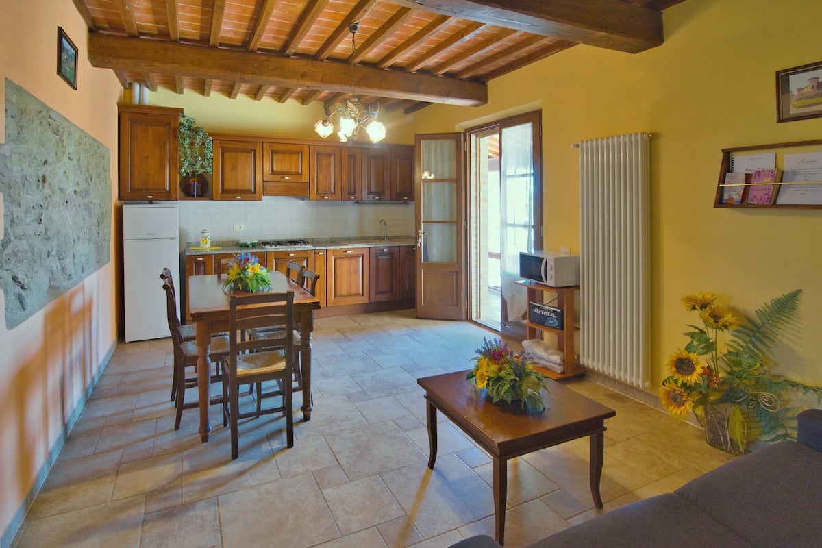 Uma sala espaçosa com uma mesa de jantar e cadeiras de madeira, uma mesa de centro e um sofá. Uma cozinha com armários de madeira e uma geladeira é visível ao fundo.