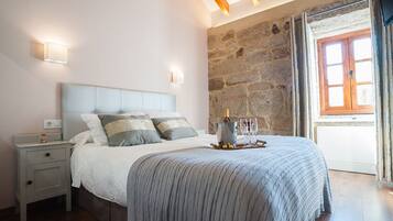 Un dormitorio con pared de piedra, techo de madera, cama con manta gris, lámparas de noche y mesita de noche.