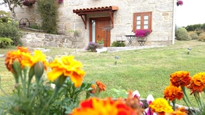 Un edificio de piedra con puerta y contraventanas de madera, un pequeño jardín con flores y un muro de piedra.