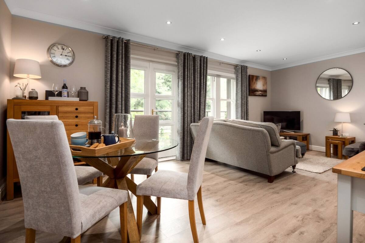 A modern living room with a glass coffee table, grey sofas, and a flat-screen TV.