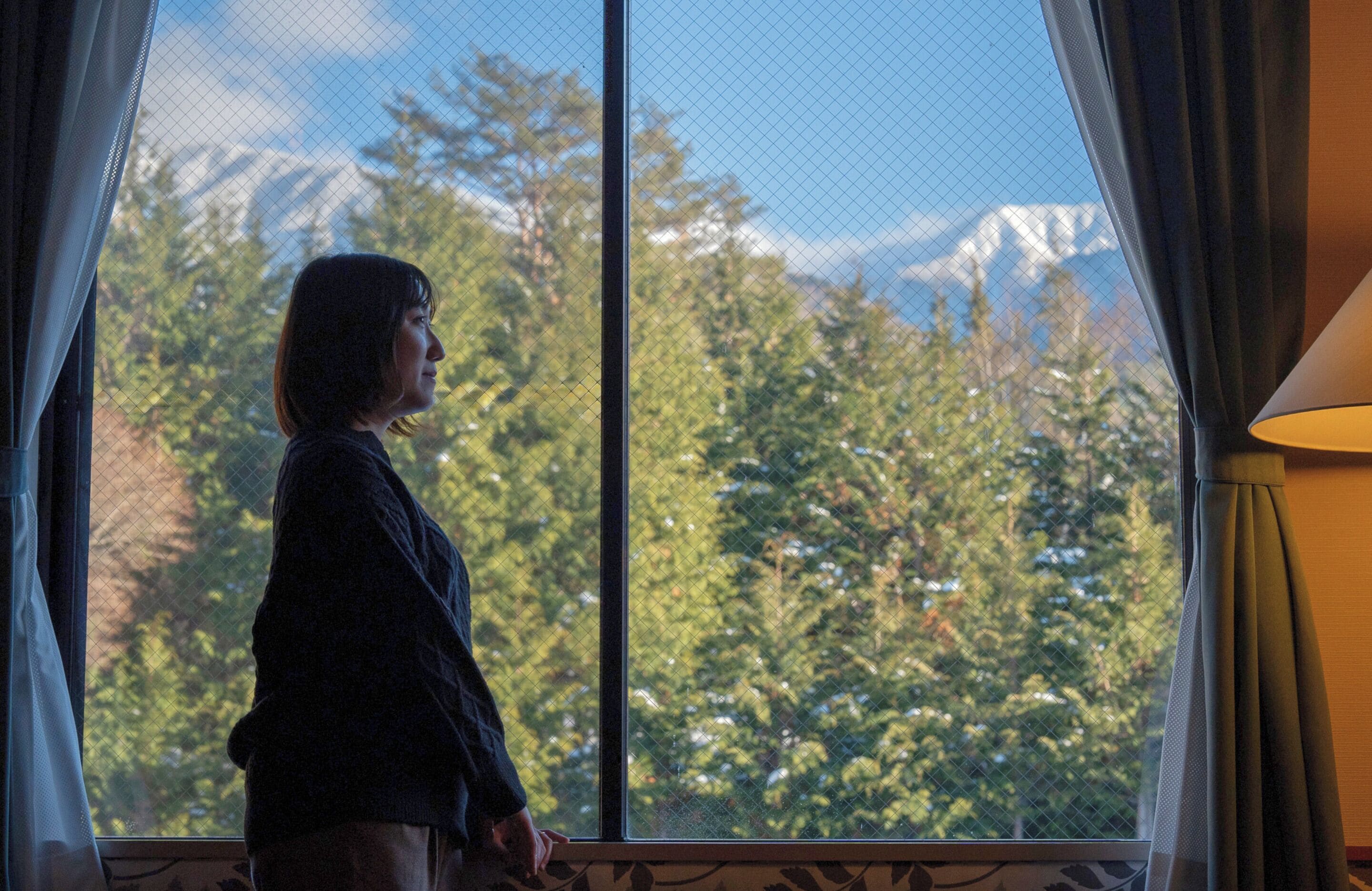 A person standing by a window with a view of a forest and mountains.