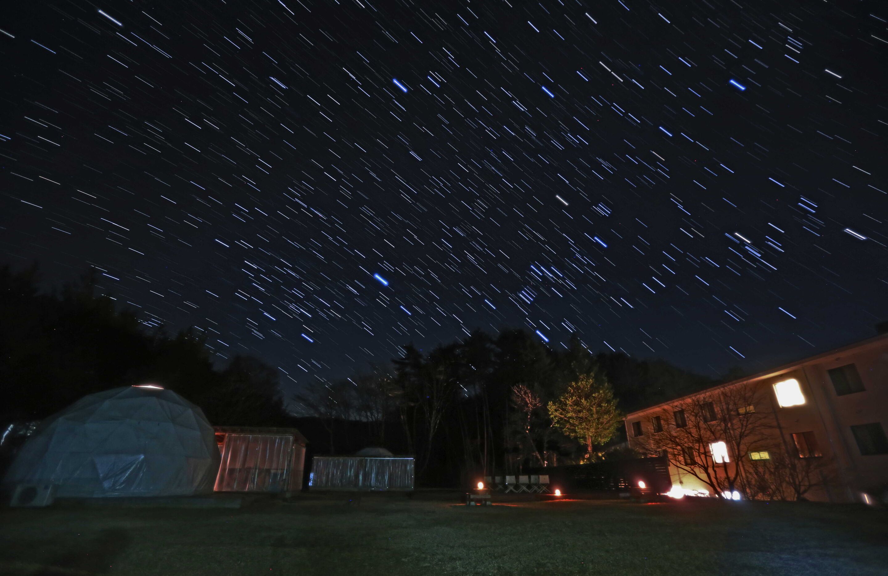 A starry night sky over a residential area with a dome-shaped structure, a building with illuminated windows, and a tree.