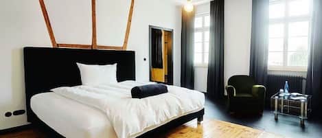 Une chambre à coucher avec une tête de lit noire, un lit recouvert de draps blancs, un plancher en bois, un fauteuil vert et une table basse en verre.