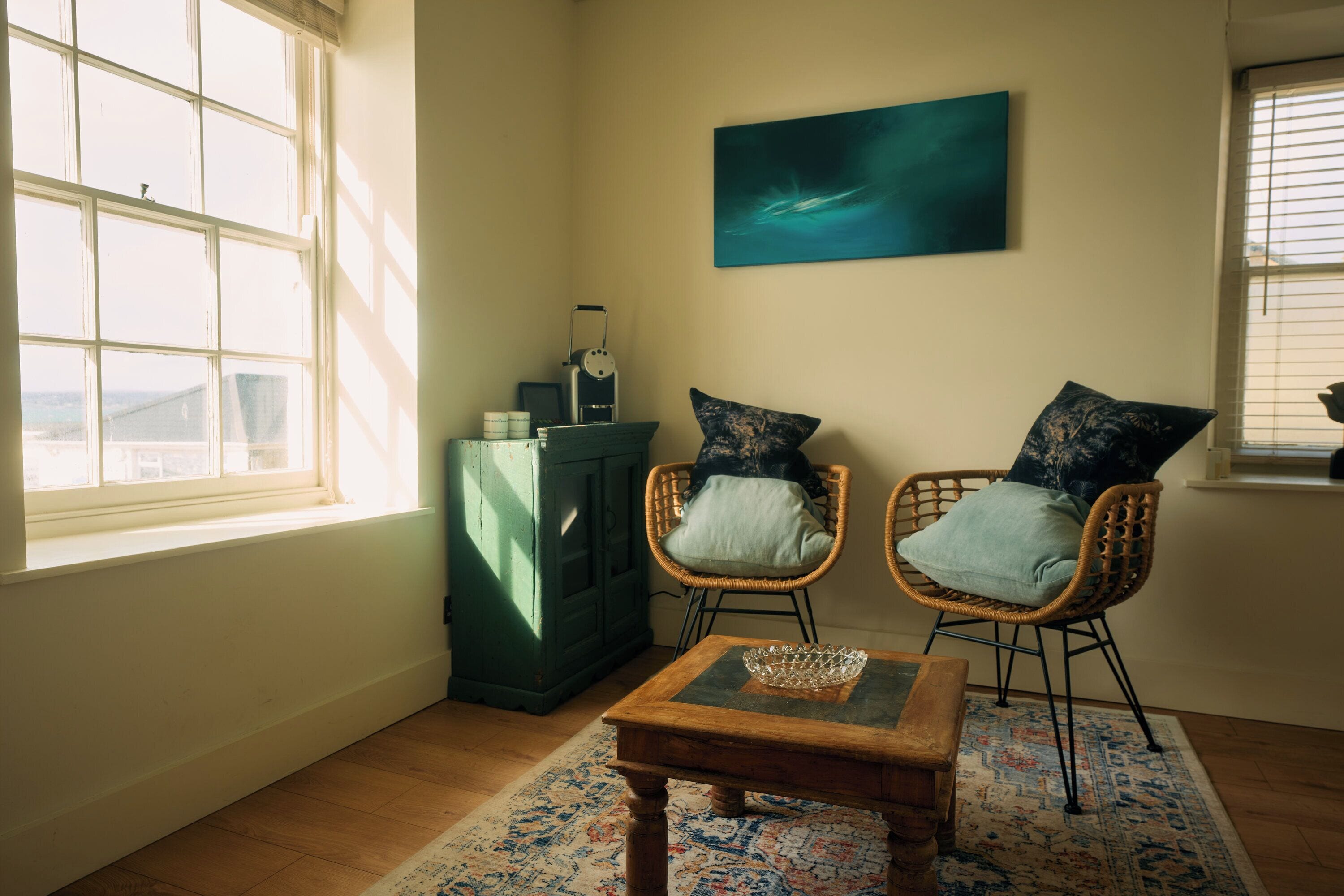 Una habitación con dos sillas de mimbre, una pequeña mesa de madera, un armario y un cuadro en la pared.