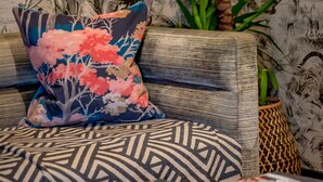 A room with a patterned wall, a sofa with patterned cushions, a decorative pillow, a small plant, and books on a table. - The Godolphin (Marazion)