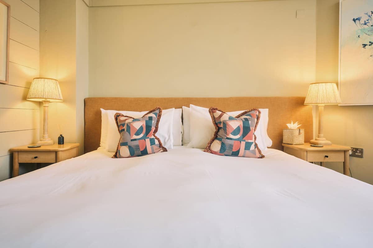 A neatly made bed with patterned pillows, two bedside lamps, a nightstand with a lamp, and a wall-mounted artwork.