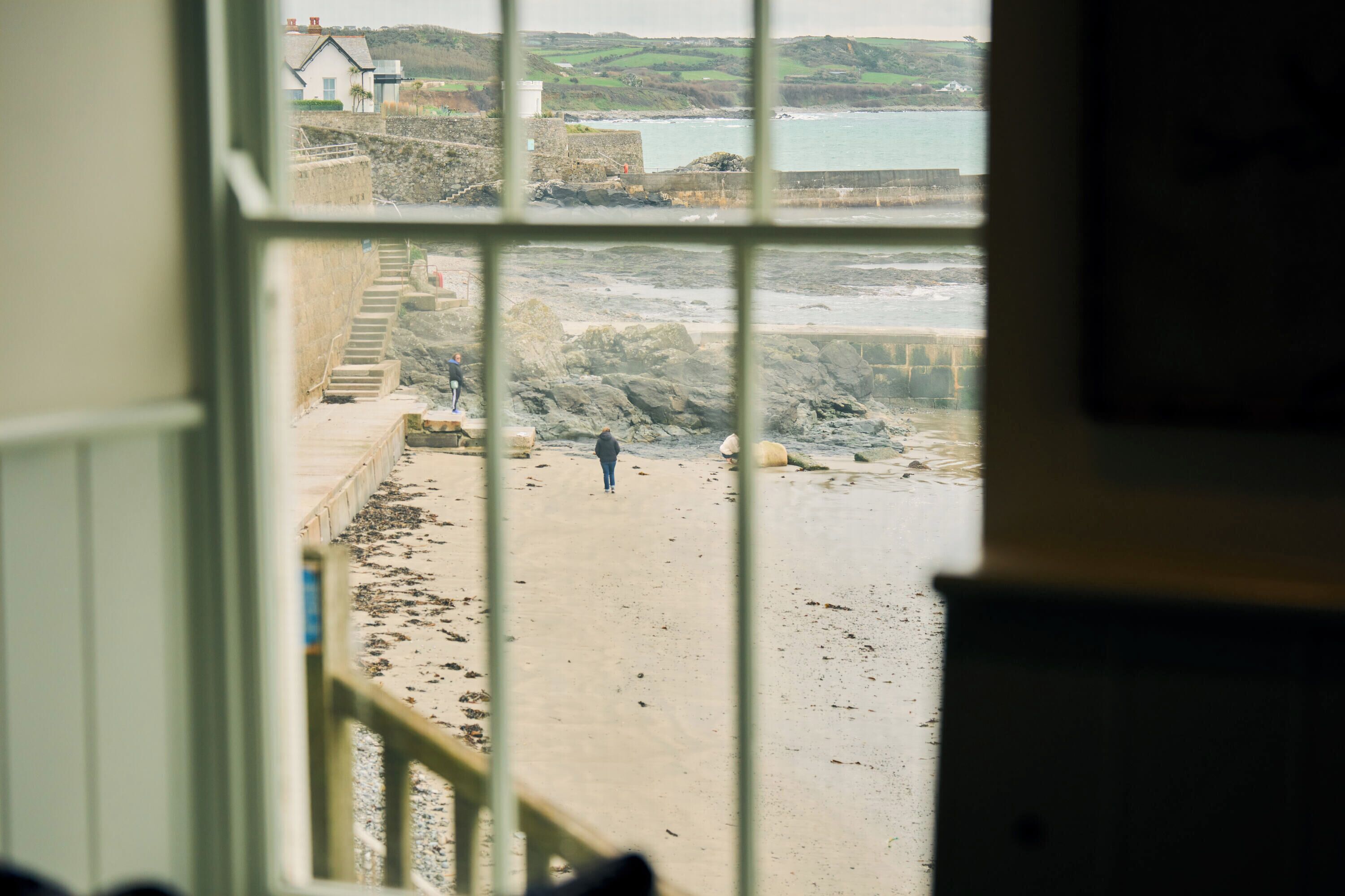 A view through a window of a sandy beach with a staircase leading down to the water, a rocky shoreline, and a building in the distance.