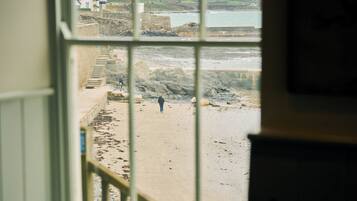 A view through a window of a sandy beach with a staircase leading down to the water, a rocky shoreline, and a building in the distance.