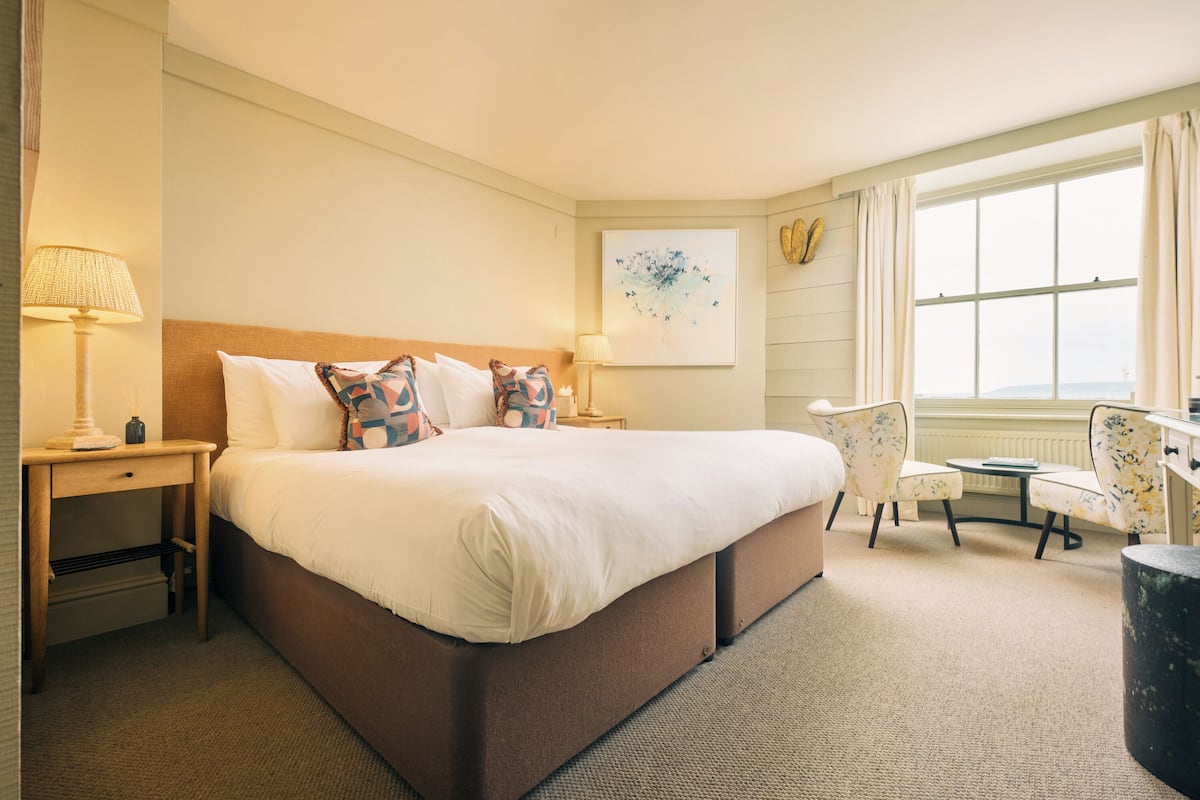 A hotel room with a large bed, bedside tables, a chair, a small table, and a window with curtains.
