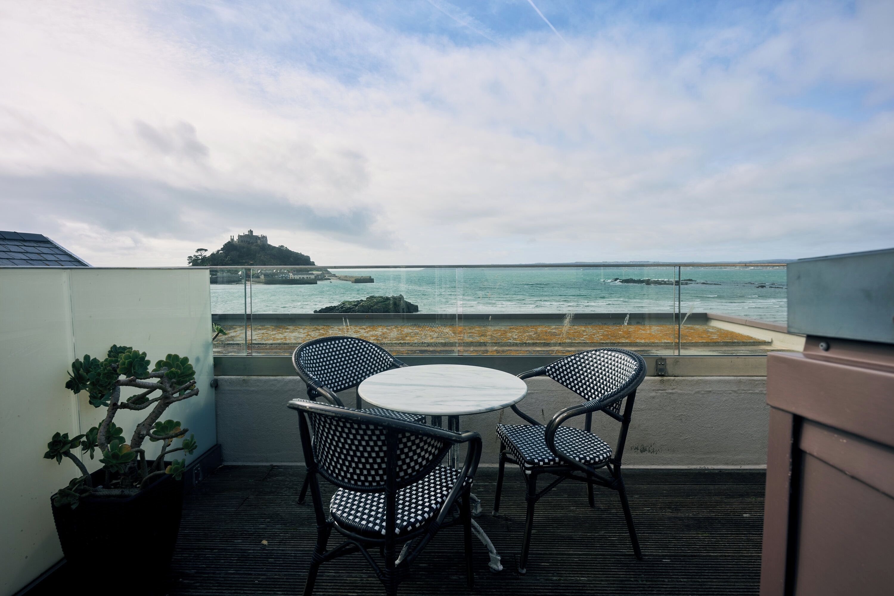 A balcony with a round table and two chairs, overlooking a coastal view with a castle on a hill and rocky outcrops in the water.